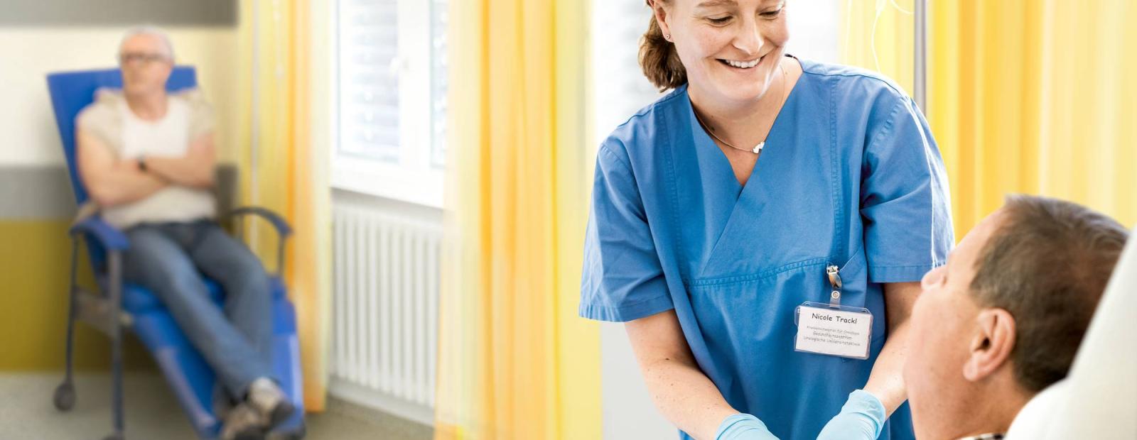 A nurse in blue scrubs smiles while attending to a seated man with an IV. Another person sits in the background in a hospital room with yellow curtains.