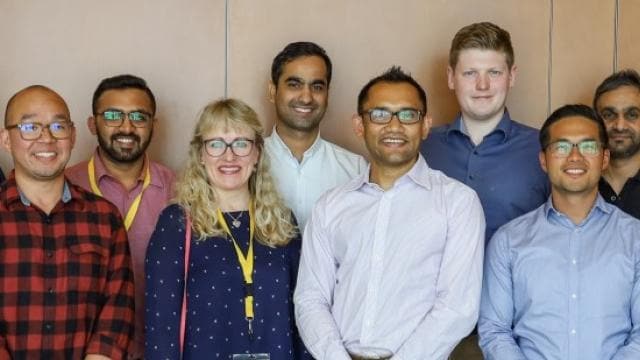A group of twelve people standing in two rows, smiling, with a wooden wall in the background.