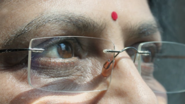 A person gazing outside, wearing glasses with a red bindi on their forehead.