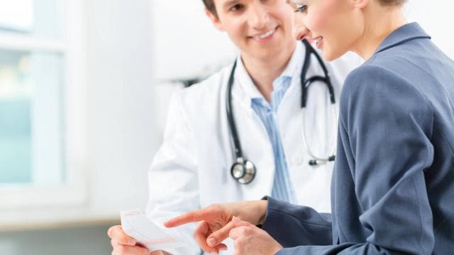 A doctor and a woman discuss documents in a medical office, with the woman pointing at a paper.