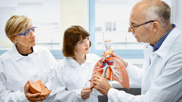 Three people in lab coats and safety glasses discuss a human respiratory system model in a laboratory setting.