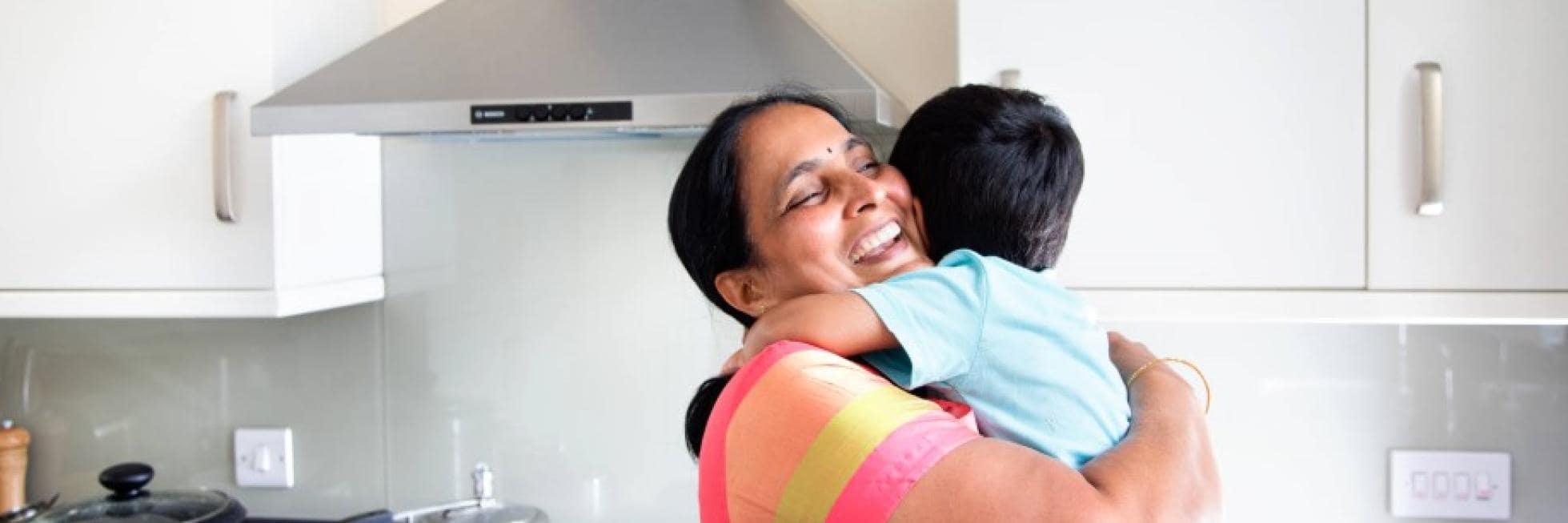 A smiling woman wearing a colorful dress embraces a young child in a kitchen.
